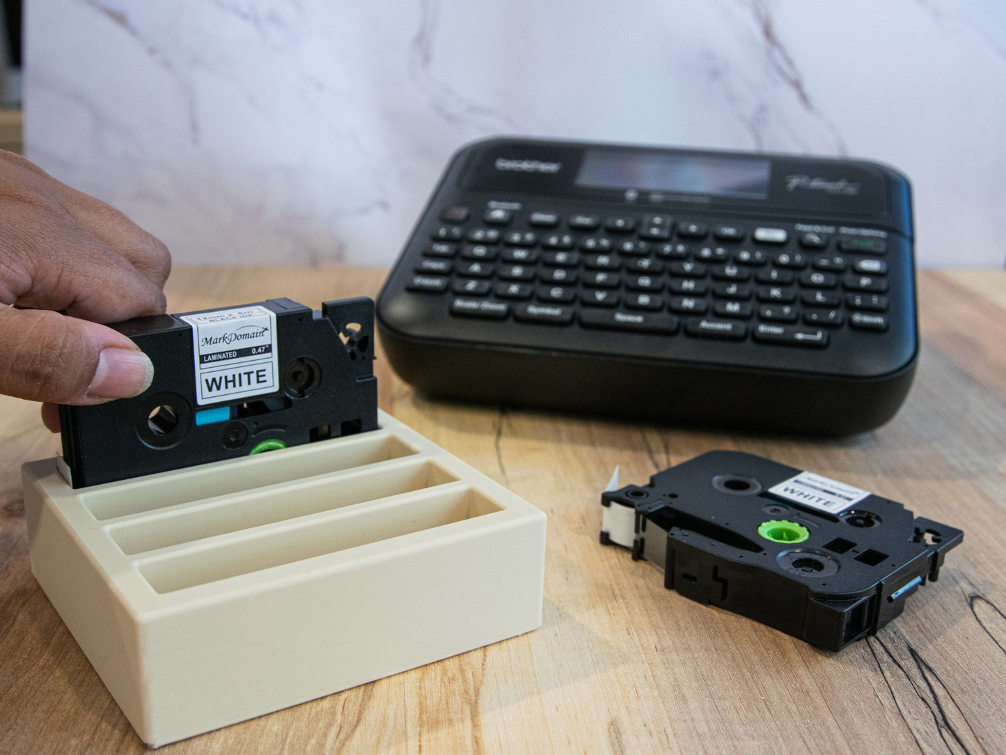 Hand placing a 12mm Brother TZe label tape into a beige organizer tray next to a Brother P-Touch label maker on a desk.