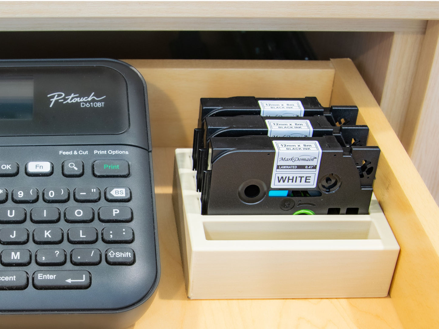 Drawer storage setup with a Brother P-Touch D610BT and a beige label tape organizer holding three 12mm TZe cartridges neatly arranged.