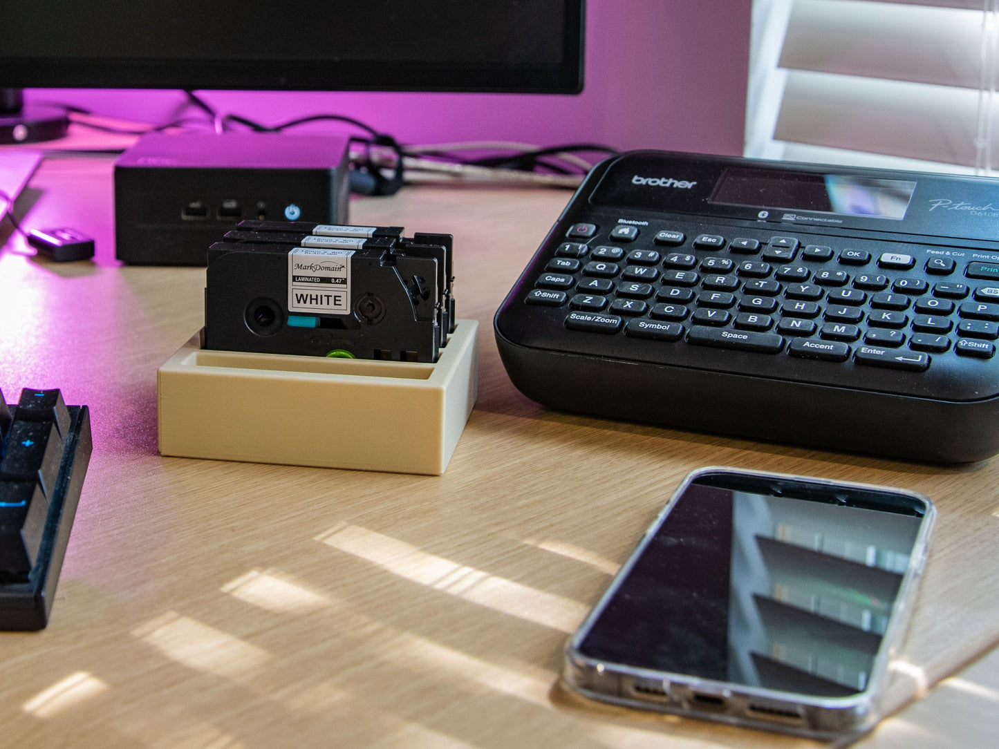 Desktop workspace featuring a Brother label maker and 12mm label tape organizer with three cartridges inserted, alongside a phone and monitor.