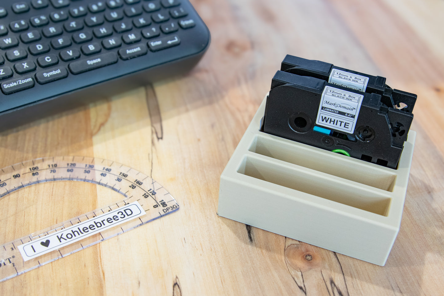 Label tape storage tray holding two Brother compatible 12mm label cartridges on a wood desk next to a keyboard and printed label reading "I ♥ Kohleebree3D".