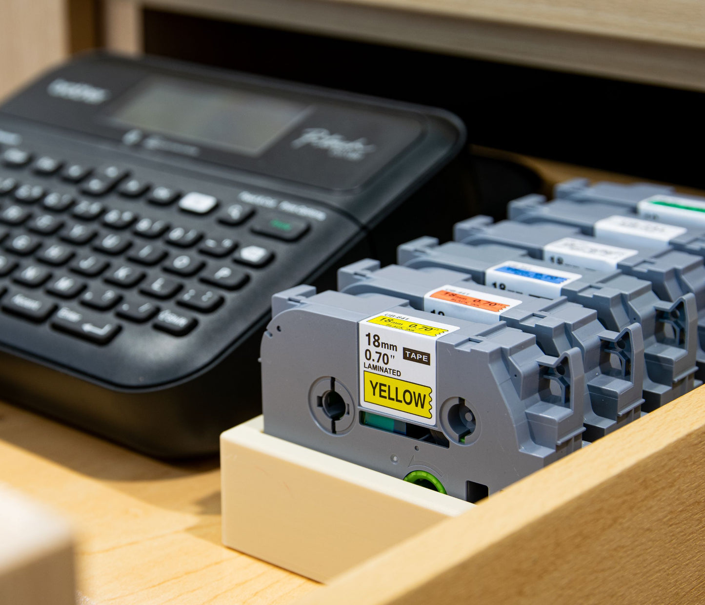 Brother P-Touch label maker next to a beige 18mm label tape organizer neatly holding colorful laminated label cartridges in a desk drawer.