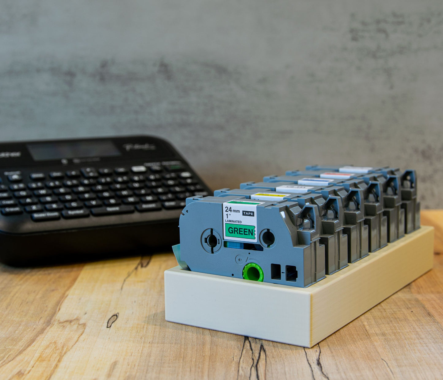 Brother label maker and 24mm tape organizer tray on a wooden desk, neatly holding multiple laminated label cartridges in a row.