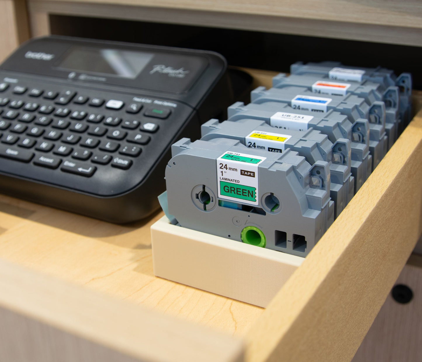 24mm Brother label tapes organized in a drawer using a beige storage tray, shown next to a P-Touch label maker for easy access and storage.