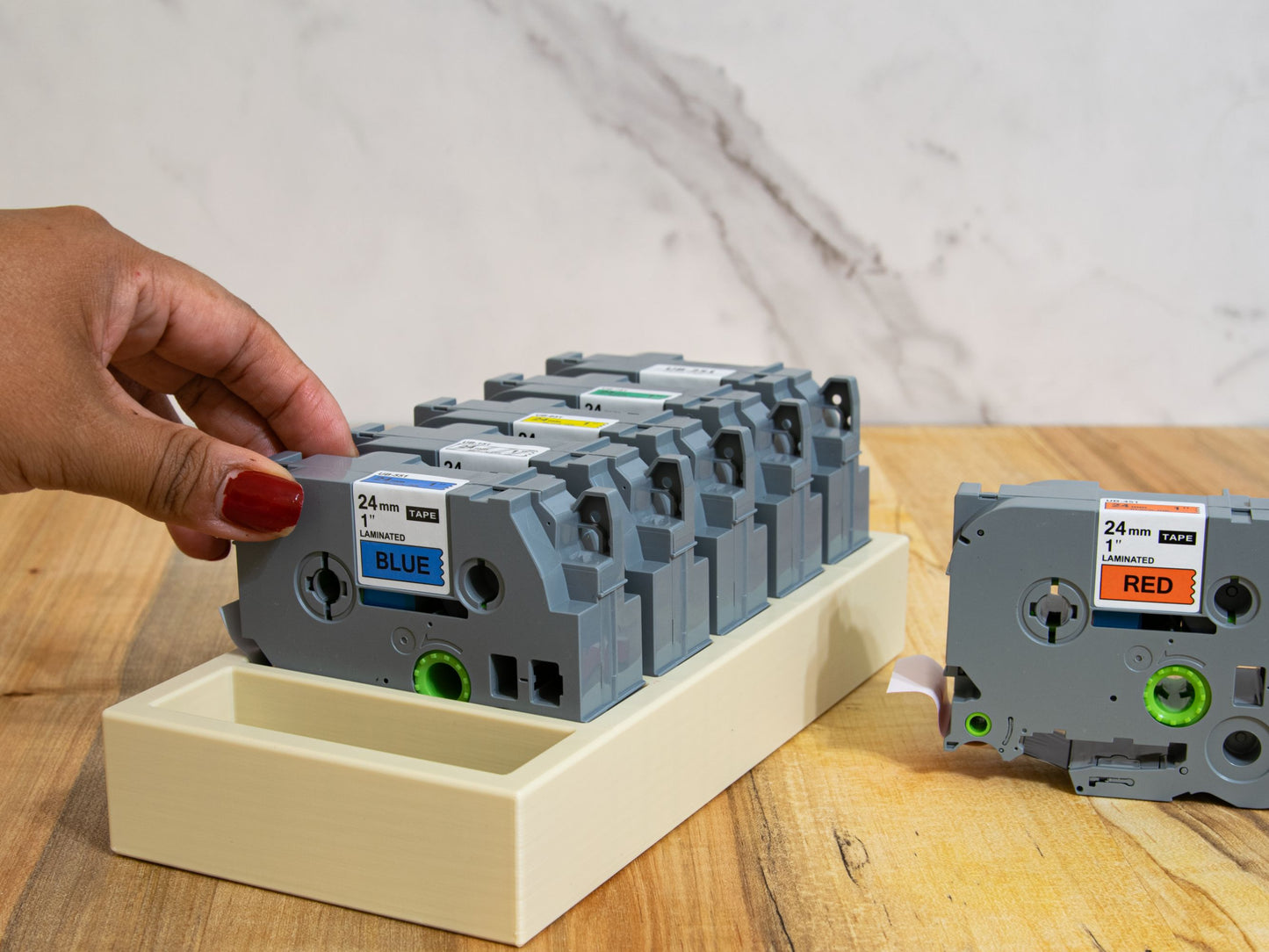 Hand removing a 24mm laminated blue label tape from a Brother-compatible organizer tray, showing ergonomic access and tidy storage solution.