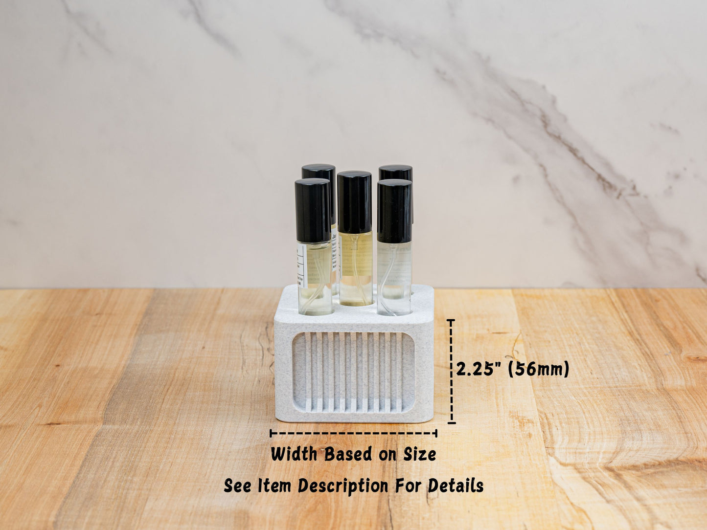 Front view of a small square perfume organizer holding four ScentSplit decant vials. It sits on a wood surface with a marble backdrop. Overlay graphics indicate the height as “2.25" (56 mm)” and note along the bottom reads: “Width Based on Brand — See Item Description For Details.” The front face features recessed vertical ribbing for a clean, architectural look.