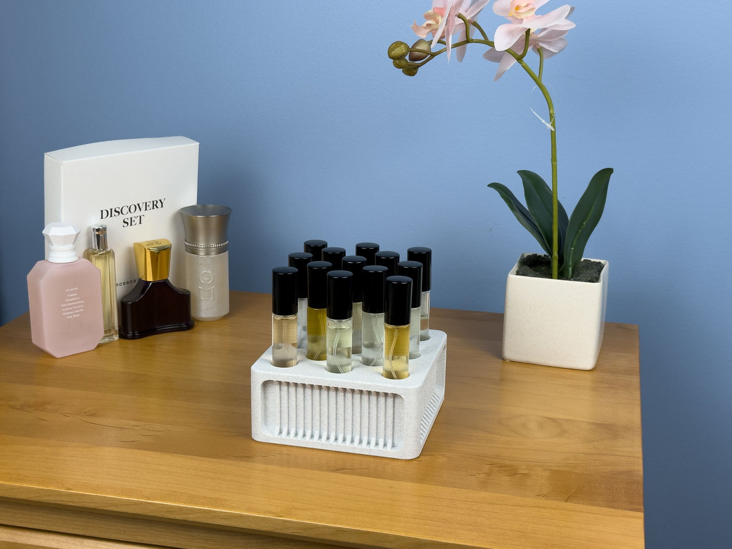 Square ScentSplit perfume organizer sits on a wooden dresser, fully loaded with ScentSplit decant vials arranged in neat rows. To the left are full-size fragrance bottles and a white “Discovery Set” box; to the right, a white planter with a pink orchid. The pale blue wall and warm wood tones create a tidy, curated vanity setup.