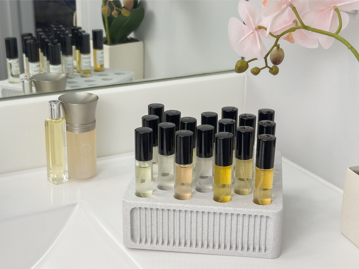 Square ScentSplit perfume organizer displayed on a white bathroom counter. Multiple ScentSplit decants are arranged, framed by a silver-trimmed container and a pink orchid flower. In the mirror reflection, another organizer is faintly visible, emphasizing its usefulness as part of a larger collection display.