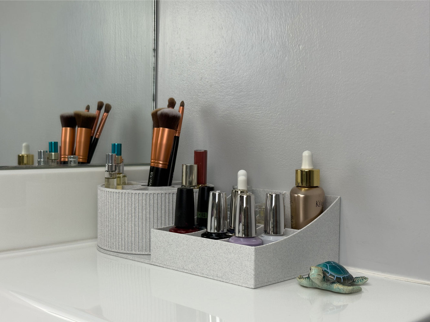 Side view of the rotating beauty organizer styled on a vanity: tall brushes and mini sprays in the rotating carousel, nail polishes and droppers arranged in the tiered base; minimalist bathroom setting with a small decorative turtle figurine for scale.