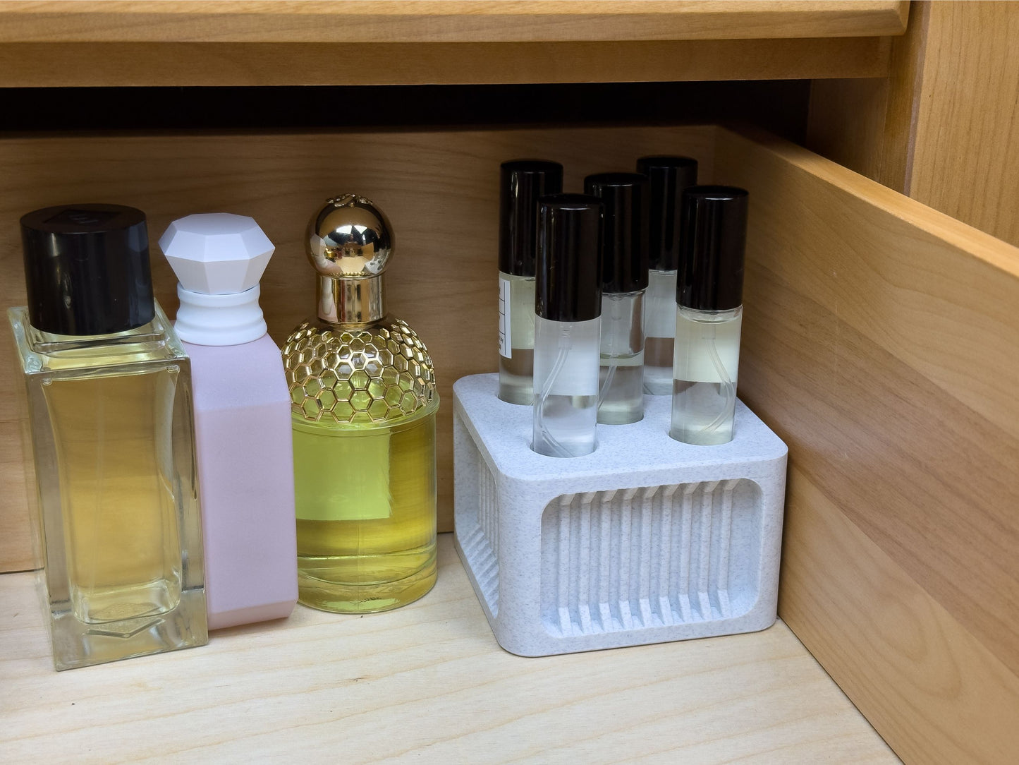 Smaller ScentSplit square perfume organizer with recessed ribbing fits neatly in the corner of a drawer, holding several ScentSplit decants upright. Around it, larger fragrance bottles in pink, gold, and clear glass are positioned side by side. The compact design shows how the organizer fits seamlessly into smaller storage areas.